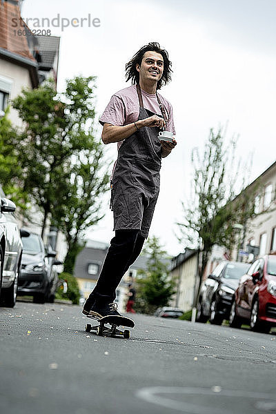 Lächelnder männlicher Barista  der eine Kaffeetasse hält  während er auf einer Straße in der Stadt Skateboard fährt
