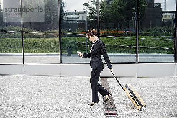 Unternehmerin  die ein Mobiltelefon benutzt  während sie mit einem Koffer auf dem Fußweg geht