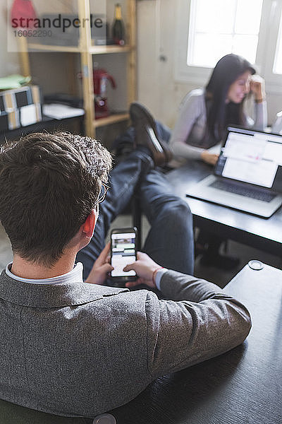 Entspannter Geschäftsmann  der sein Smartphone am Schreibtisch benutzt  während eine Frau im Hintergrund arbeitet