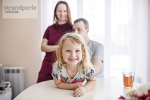 Glückliches süßes Mädchen lehnt sich an Esstisch mit Eltern stehen im Hintergrund zu Hause