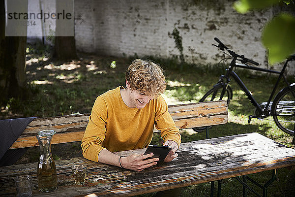 Lachender junger Mann sitzt am Biertisch im Garten und benutzt ein digitales Tablet