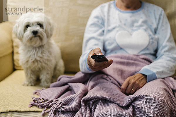 Kranke Frau im Ruhestand sitzt mit ihrem Hund auf dem Sofa und sieht zu Hause fern