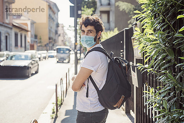 Junger Mann mit Maske und Rucksack steht auf dem Bürgersteig in der Stadt