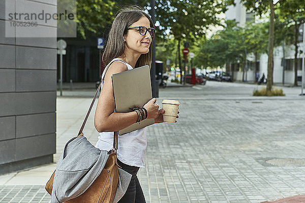 Nachdenkliche Geschäftsfrau mit Laptop und Einwegglas beim Spaziergang auf der Straße