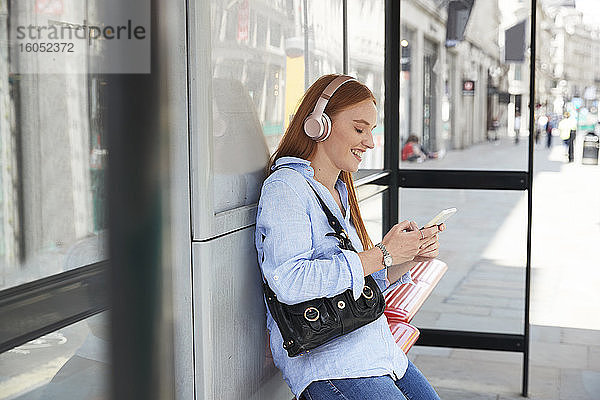 Lächelnde junge Frau  die Musik hört und ihr Smartphone an der Bushaltestelle benutzt