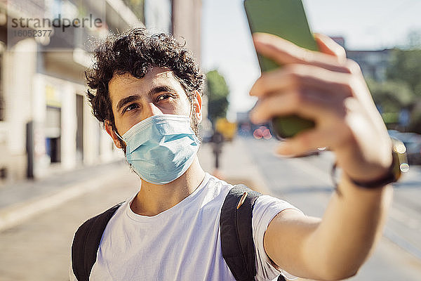 Nahaufnahme eines Mannes mit Gesichtsmaske  der ein Selfie mit einem Smartphone in der Stadt macht