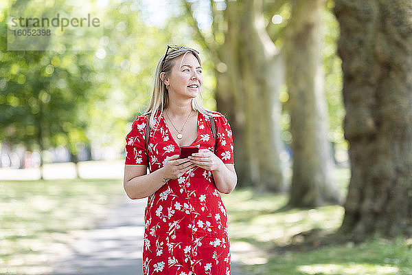 Nachdenkliche Frau  die ihr Smartphone in der Hand hält  während sie auf dem Fußweg in einem öffentlichen Park spazieren geht