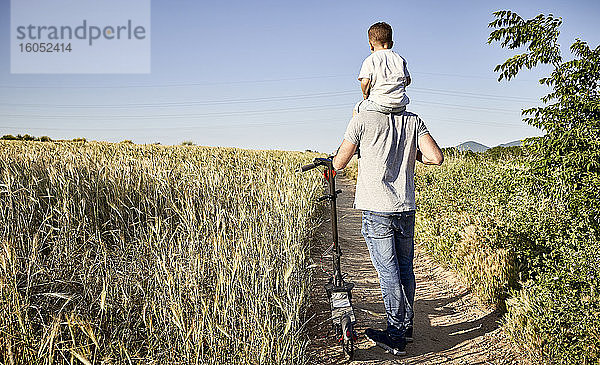 Vater  der seinen Sohn auf den Schultern trägt  während er mit dem Tretroller inmitten von Pflanzen spazieren geht