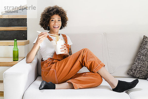 Porträt einer glücklichen jungen Frau  die zu Hause auf der Couch sitzt und asiatisches Essen isst