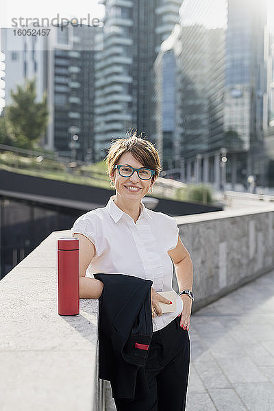 Lächelnde Geschäftsfrau mit Brille an einer Stützmauer in der Stadt