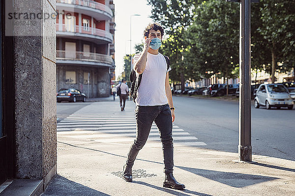 Mann mit Gesichtsmaske  der über sein Smartphone spricht  während er in der Stadt auf der Straße geht