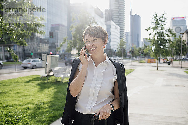 Lächelnde Geschäftsfrau  die über ein Smartphone spricht und wegschaut  während sie auf dem Bürgersteig in der Stadt steht