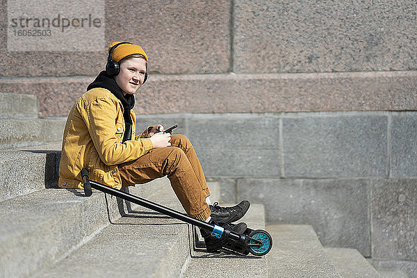 Nachdenklicher Junge hört Musik  während er mit seinem Roller auf einer Treppe sitzt  während eines sonnigen Tages