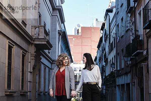 Lesbisches Paar hält sich an den Händen  während es inmitten von Gebäuden in der Stadt spazieren geht
