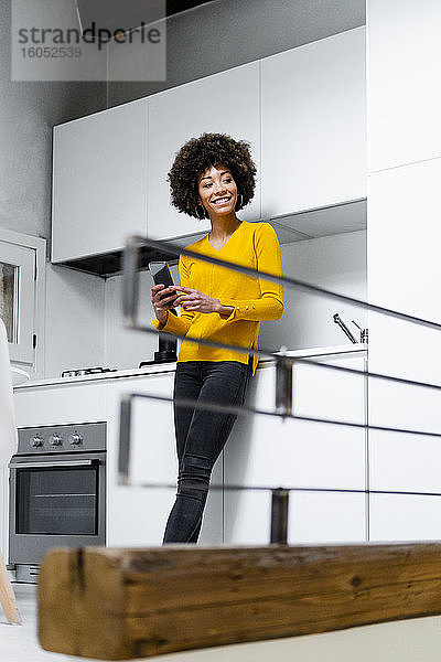Porträt einer lächelnden jungen Frau  die mit einem Smartphone in der Küche steht