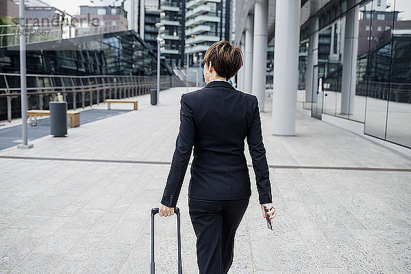 Geschäftsfrau mit kurzen Haaren  die einen Koffer hält  während sie auf einem Fußweg in der Stadt spazieren geht