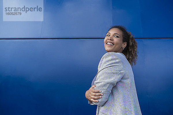Lächelnde Geschäftsfrau mit verschränkten Armen vor einer blauen Wand