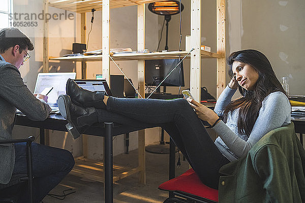 Entspannte Frau mit Smartphone am Schreibtisch mit Geschäftsmann mit Laptop