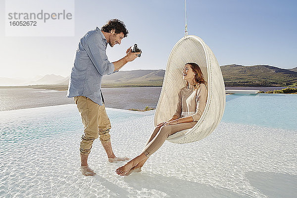 Mann fotografiert Frau  die in einem Hängesessel über dem Schwimmbad sitzt