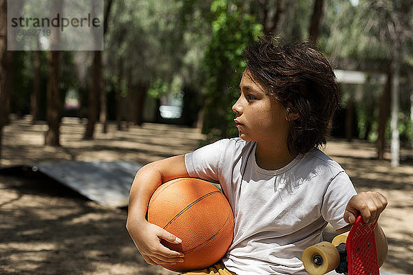 Nachdenklicher Junge mit Basketball und Skateboard im Park
