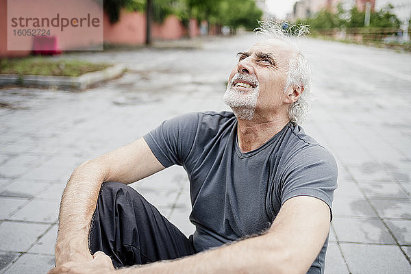 Müder älterer Mann schaut auf  während er auf einem Fußweg in der Stadt sitzt