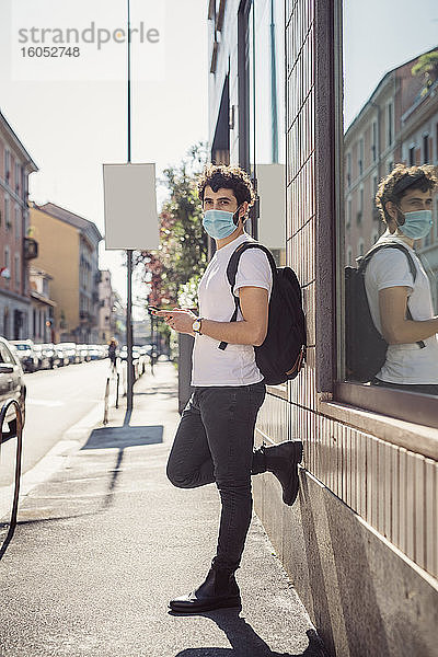 Mann mit Maske  der ein Smartphone benutzt  während er an einem Gebäude in der Stadt steht