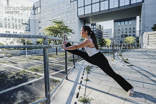 Junge sportliche Frau streckt die Beine auf einem Geländer in der Stadt an einem sonnigen Tag