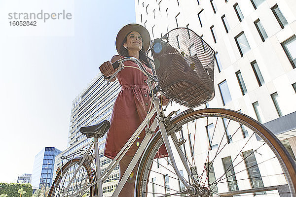 Nachdenkliche Frau steht mit Fahrrad gegen Gebäude und Himmel an einem sonnigen Tag