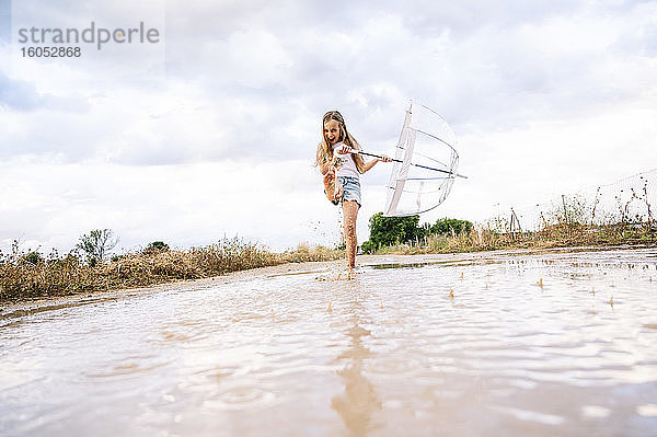 Fröhliches Mädchen spritzt Wasser in einer Pfütze und hält einen Regenschirm gegen den bewölkten Himmel