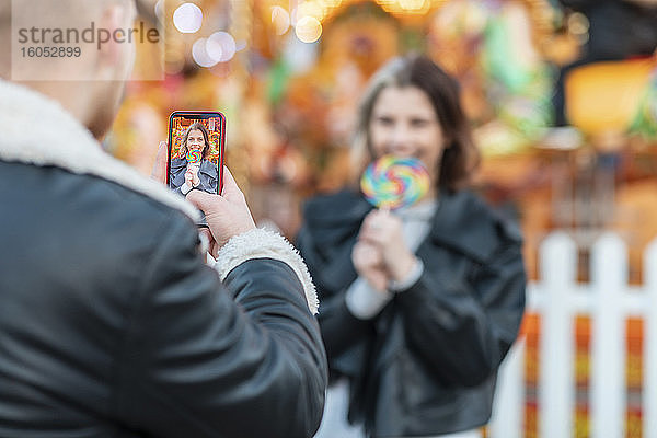 Mann fotografiert Freundin beim Essen eines Lutschers mit dem Smartphone im Vergnügungspark