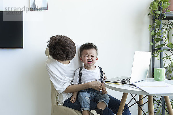 Mutter hält weinenden Jungen auf dem Schoß  während sie über einem Stuhl zu Hause sitzt