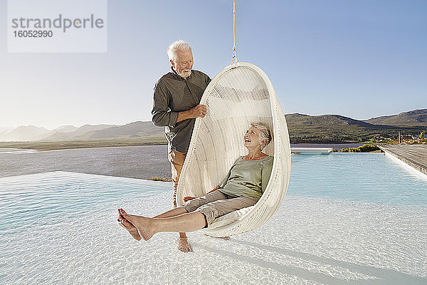 Glückliches älteres Paar mit Frau  die in einem Hängesessel über dem Schwimmbad sitzt