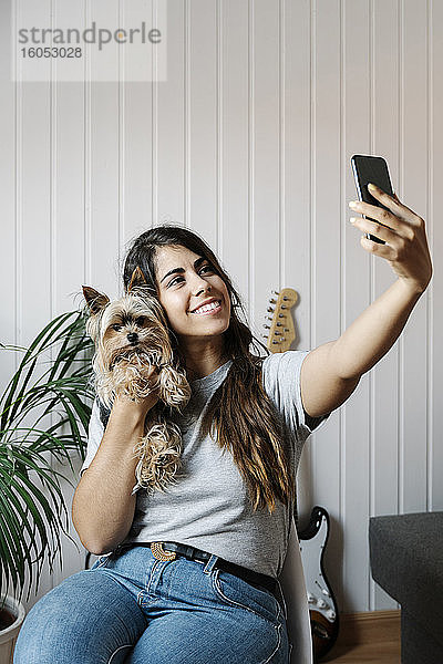 Junge Frau macht Selfie mit Yorkshire Terrier  während sie zu Hause auf einem Stuhl sitzt