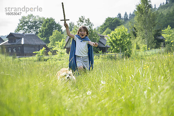 Verspielter Junge mit Umhang läuft mit Hund auf einer Wiese