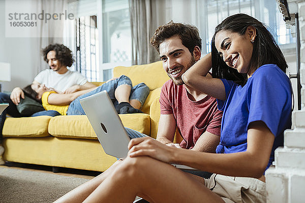 Lächelndes junges Paar  das sich einen Laptop teilt  während sich Freunde auf dem Sofa im Wohnzimmer entspannen