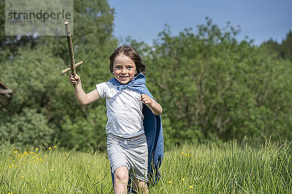 Verspielter Junge mit Umhang  der ein Spielzeugschwert hält  während er auf einer Wiese läuft