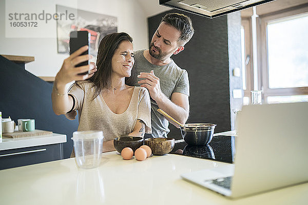 Verspielter Mann  der Mehl auf das Gesicht einer Frau aufträgt  während sie ein Selfie mit einem Smartphone zu Hause macht