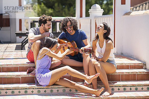 Fröhliche junge Freunde  die mit einer Gitarre auf den Stufen einer Terrasse sitzen und sich an einem sonnigen Tag vergnügen