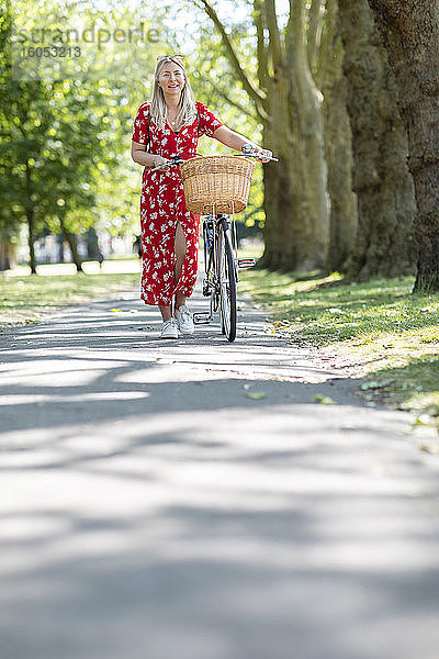 Glückliche Frau  die mit dem Fahrrad auf einem Fußweg in einem öffentlichen Park an einem sonnigen Tag spazieren geht