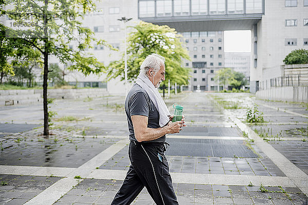 Älterer Mann  der eine Wasserflasche hält und Musik hört  während er auf einem Fußweg in der Stadt spazieren geht