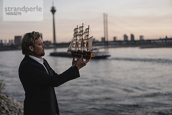 Geschäftsmann hält Modellschiff am Flussufer in der Abenddämmerung