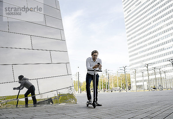 Geschäftsmann mit Kick-Scooter und Smartphone spiegelt sich in Gebäudefassade