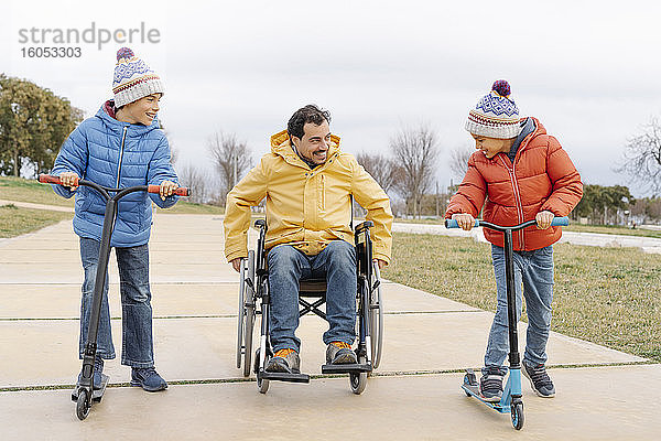 Fröhliche Jungen fahren Roller  während der Vater im Rollstuhl auf dem Gehweg im Park sitzt