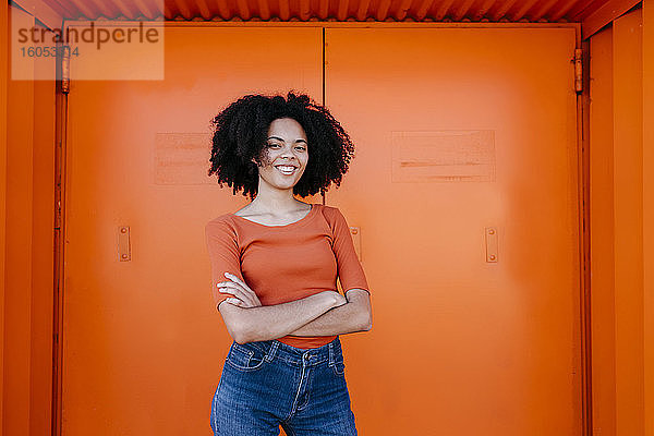 Lächelnde junge Afro-Frau steht mit verschränkten Armen vor einer orangefarbenen Tür