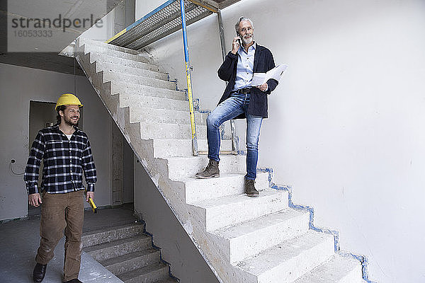 Männlicher Architekt  der über sein Smartphone spricht  während ein Bauarbeiter in einem im Bau befindlichen Haus arbeitet
