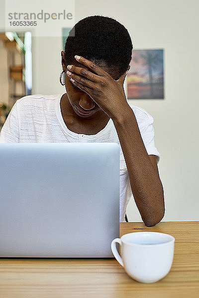 Junge Frau mit Kopf in der Hand  die einen Laptop auf einem Tisch zu Hause benutzt
