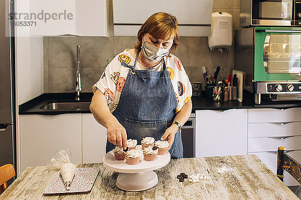 Weibliche Bäckerin mit Maske beim Verzieren von Törtchen auf einem Tortenständer in einer Werkstatt