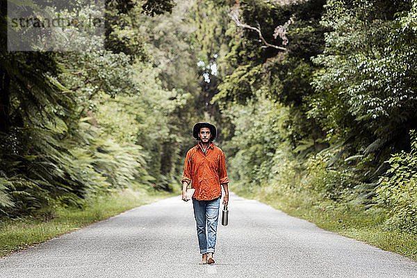 Selbstbewusster junger Mann  der eine Wasserflasche hält  während er auf einer Straße im Wald spazieren geht