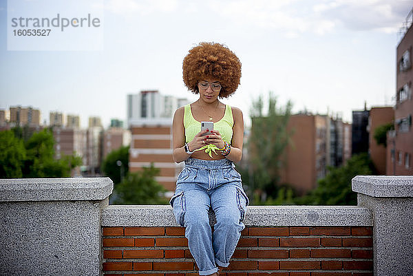 Frau mit Afrofrisur  die ihr Smartphone benutzt  während sie auf einer Stützmauer gegen den Himmel sitzt