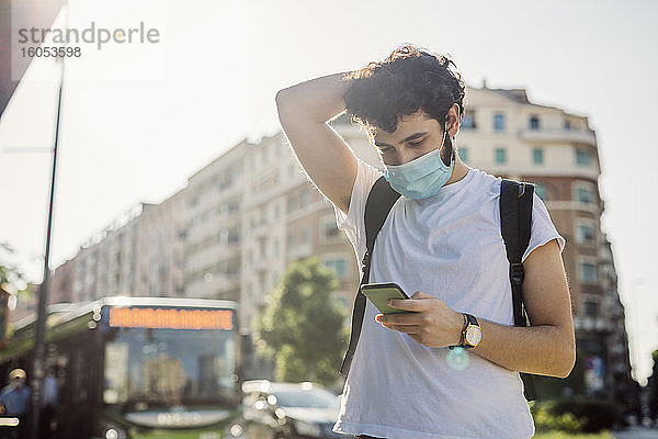 Junger Mann mit Gesichtsmaske  der sein Smartphone benutzt  während er an einem sonnigen Tag in der Stadt steht